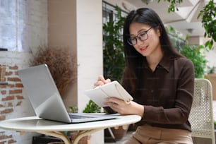 Smiling businesswoman sitting at outdoor and working with computer laptop and looking information on notebook.