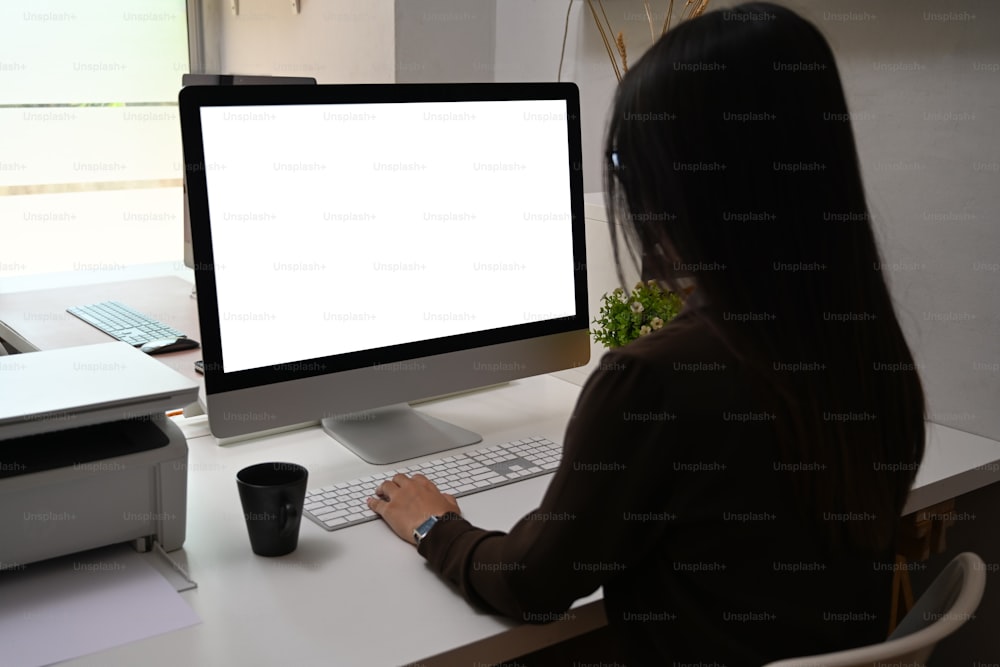 Rückansicht einer jungen Mitarbeiterin, die mit dem Computer arbeitet, während sie an einem modernen Arbeitsplatz sitzt.