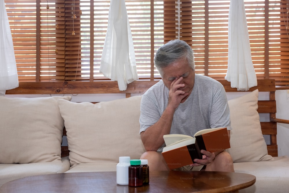 Homem sênior asiático sentado no sofá na sala de estar de casa e lendo um livro. Avô idoso estressado que sofre de fadiga ocular e dor de cabeça. Aposentadoria idosos cuidados de saúde e conceito de problema de visão ocular.