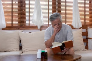 Asian senior man sitting on sofa in home living room and reading a book. Stressed elderly grandfather suffering from eye strain and headache. Retirement old people health care and eye vision problem concept.