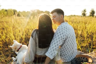 Elegante y hermosa pareja con perro blanco que se relaja en una manta en una luz cálida y soleada entre la hierba en el prado de verano. Vacaciones de verano y picnic. Familia joven disfrutando de la puesta del sol con el cachorro de pastor suizo