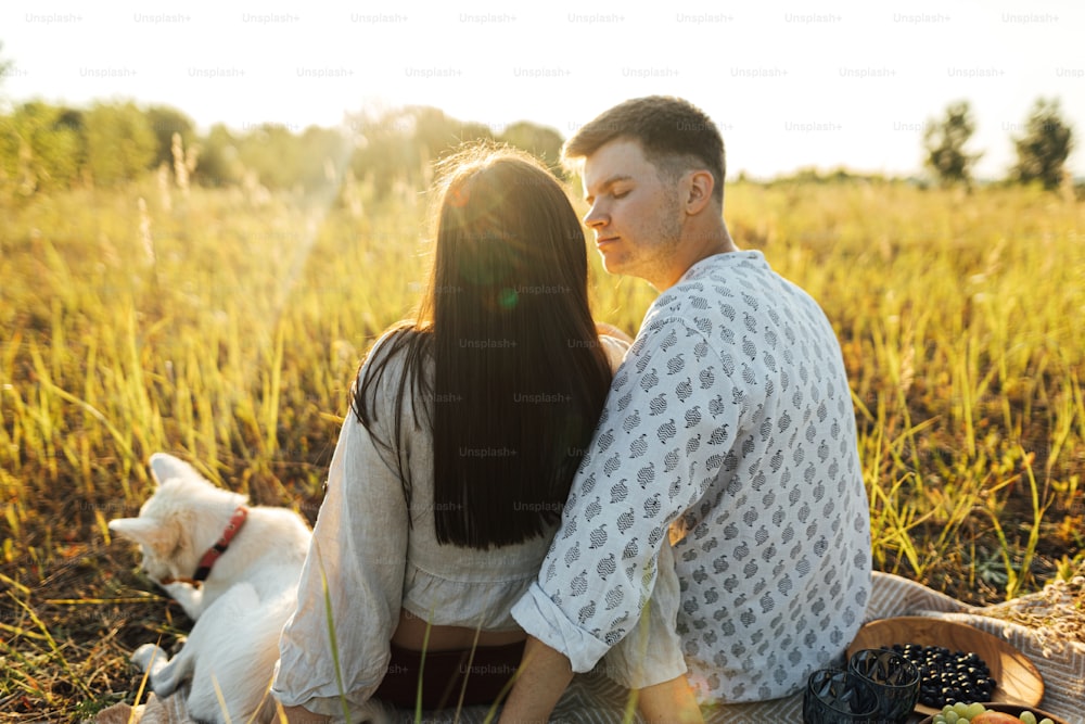 Elegante casal bonito com cão branco relaxando no cobertor em luz quente e ensolarada entre a grama no prado de verão. Férias de verão e piquenique. Família jovem que aprecia o pôr do sol com filhote de pastor suíço