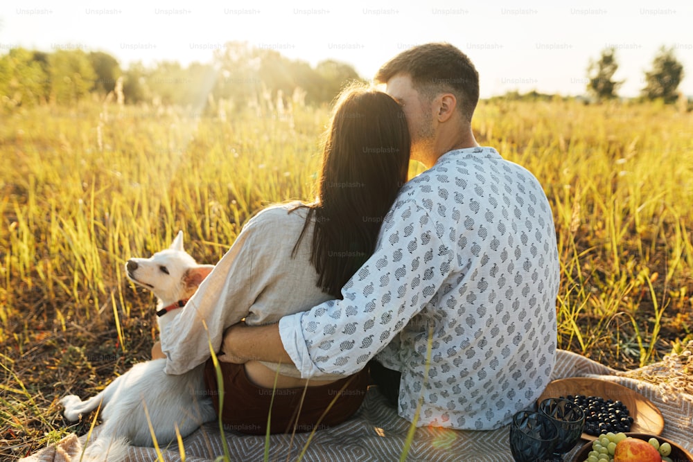 Stylish beautiful couple with white dog relaxing on blanket in warm sunny light among grass in summer meadow. Summer vacation and picnic. Young family enjoying sunset with swiss shepherd puppy