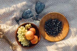 Picnic estivo. Frutta fresca deliziosa in piatti di legno e bicchieri da vino blu su coperta gialla in una calda luce soleggiata nel prato estivo. Vacanze e tempo libero