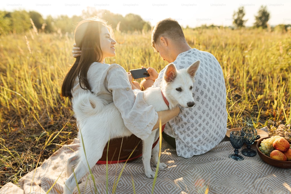 Stilvolles schönes Paar mit weißem Hund, das sich auf einer Decke in warmem, sonnigem Licht zwischen Gras auf der Sommerwiese entspannt. Sommerurlaub und Picknick. Junge Familie genießt den Sonnenuntergang mit Schweizer Schäferhund Welpe