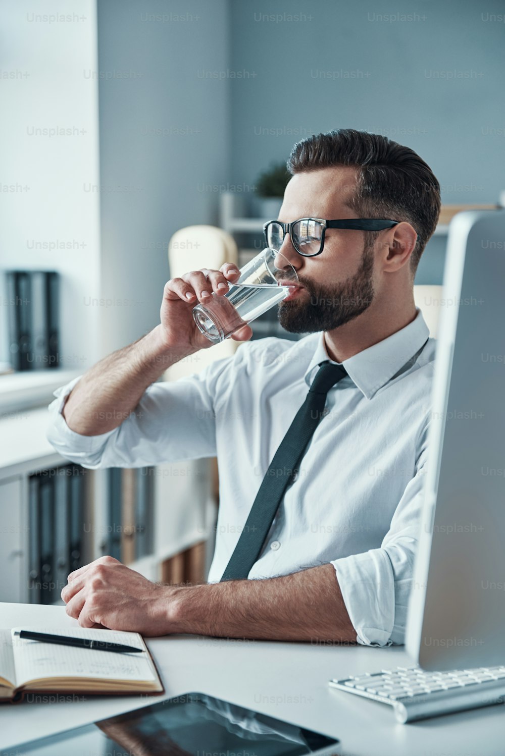 Jovem bonito de camisa e gravata bebendo água enquanto está sentado no escritório