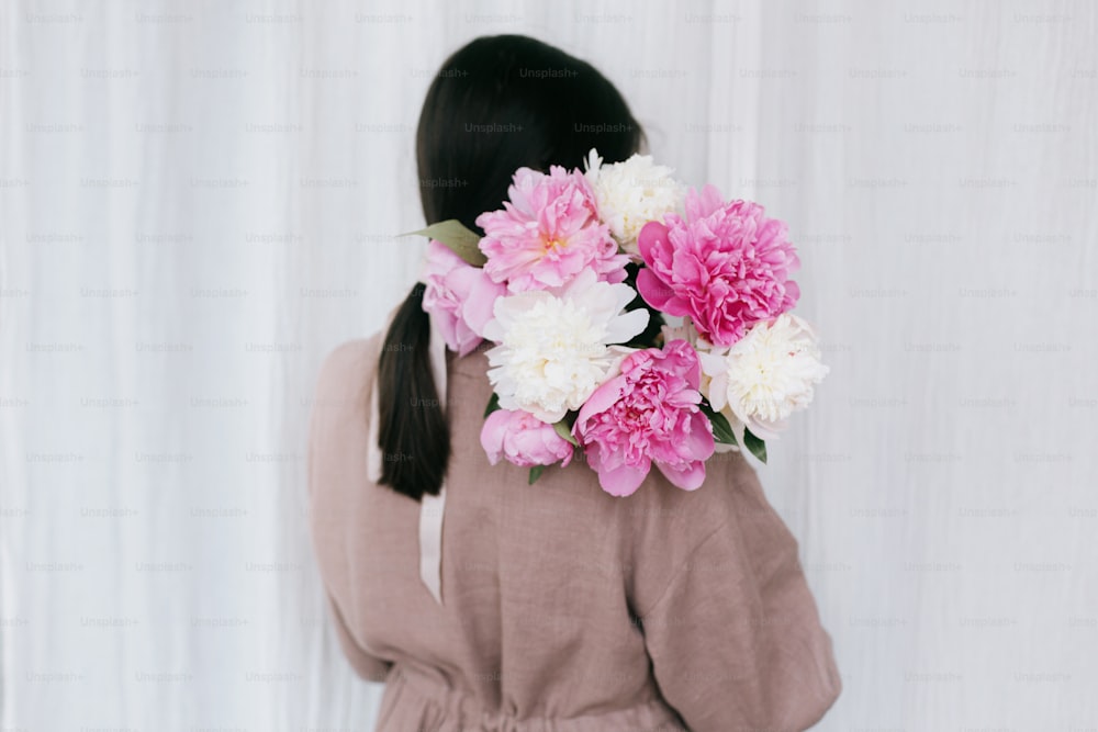 Stylish woman in linen dress holding peony bouquet on background of white fabric. Slow life, little happy things. Young female in boho rustic dress hiding face with pink and white peonies.