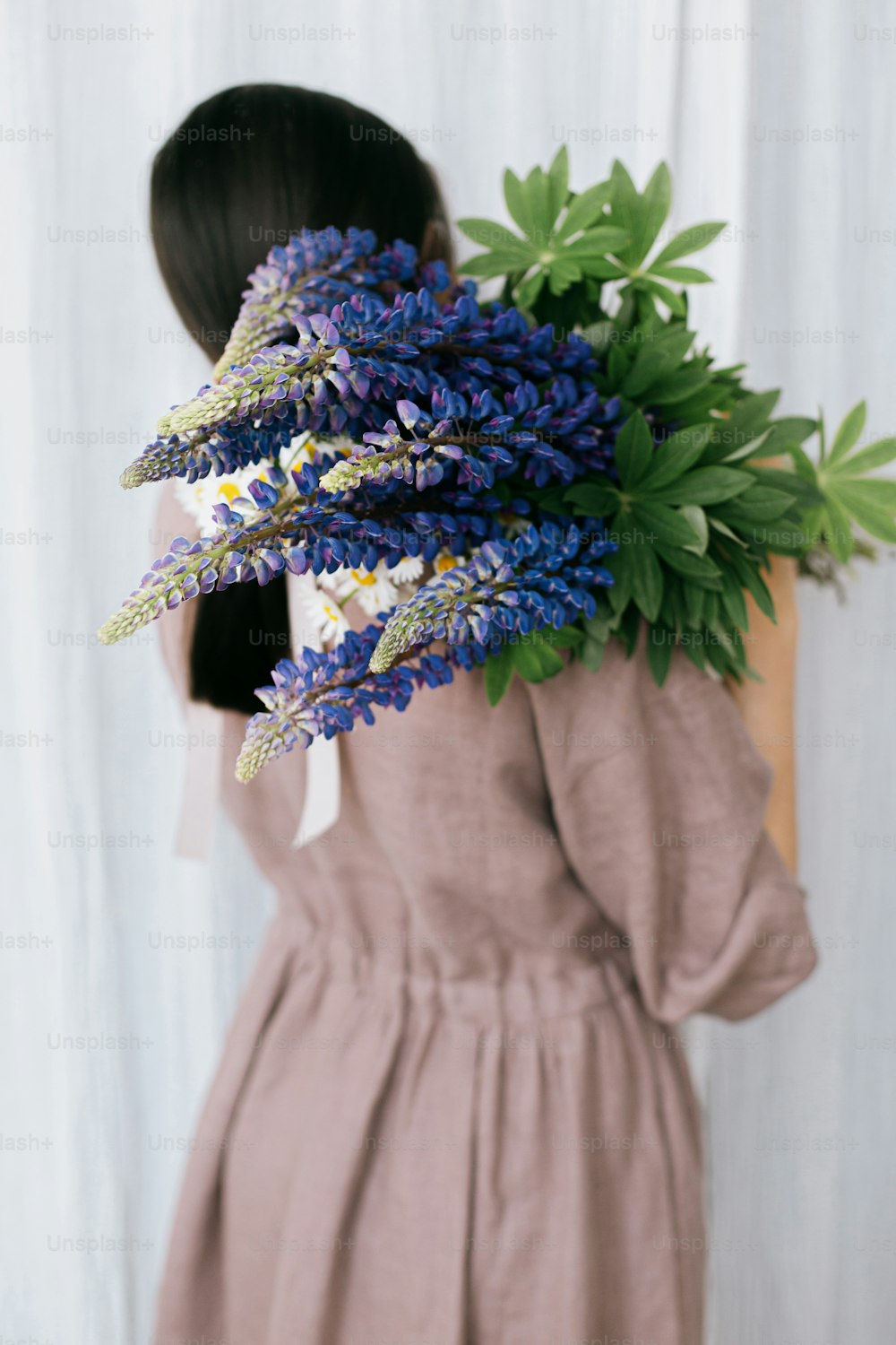 Stylish woman in linen dress holding lupine bouquet on background of pastel fabric. Simple slow living. Young female in boho rustic dress hiding face with lupine and daisy wildflowers. Aesthetics