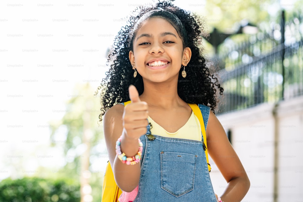 アフリカ系アメリカ人の小さな女の子は、路上でバックパックを背負いながら、笑顔で親指を立てています。教育のコンセプト。