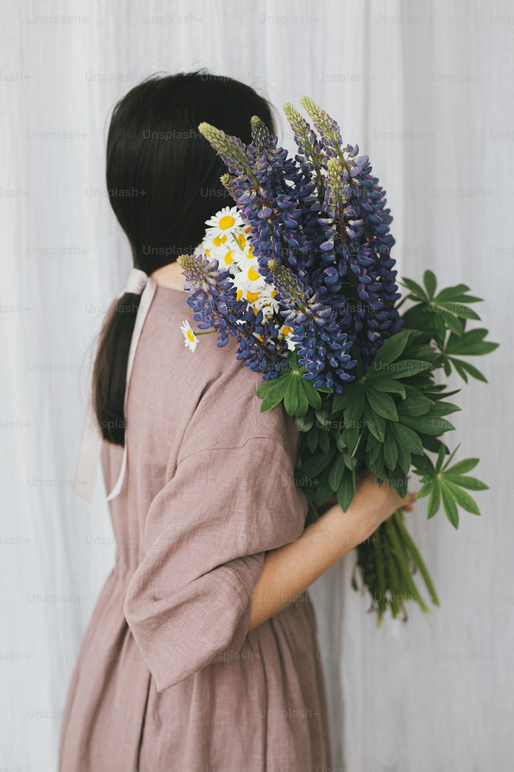 Stylish woman in linen dress holding lupine bouquet on background of pastel fabric. Simple slow living. Young female in boho rustic dress hiding face with lupine and daisy wildflowers. Aesthetics
