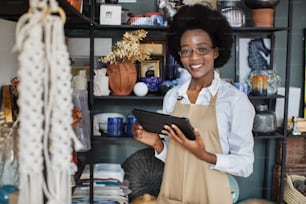 Close up of smiling pretty afro american woman in eyegasses using digital tablet for making inventory at decor shop. Attractive saleswoman in apron standing with modern gadget in hands.