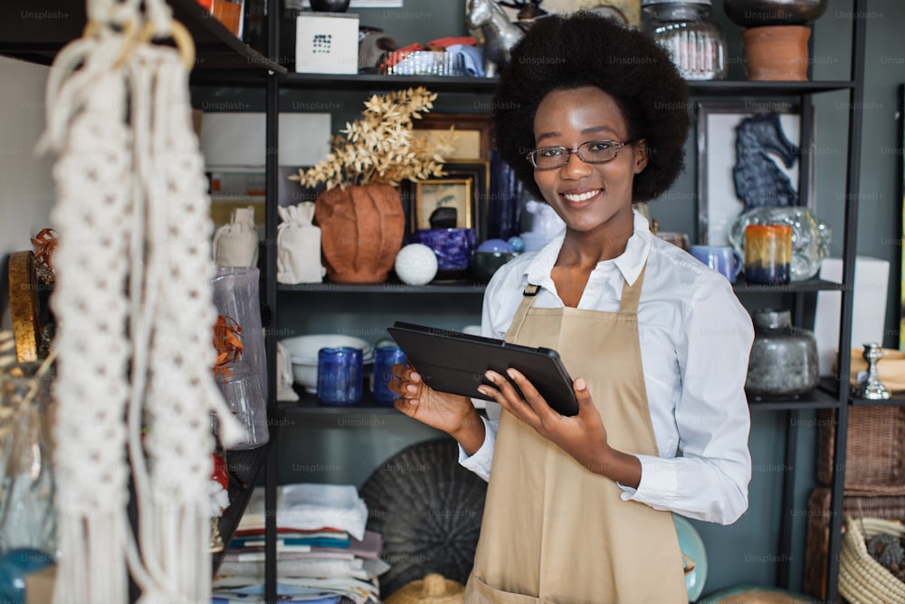 Primo piano di una bella donna afroamericana sorridente in eyegasses che utilizza la tavoletta digitale per fare l'inventario nel negozio di decorazioni. Commessa attraente in grembiule in piedi con gadget moderno in mano.