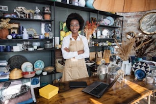 Portrait d’une sympathique femme afro-américaine en uniforme et lunettes souriant et regardant la caméra. Boutique moderne avec divers décors sur les étagères.