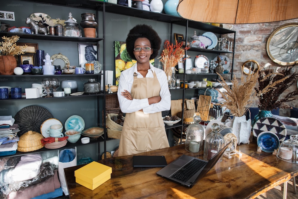 Portrait of likable afro american woman in uniform and eyeglasses smiling and looking at camera. Modern shop with various decor on shelves.