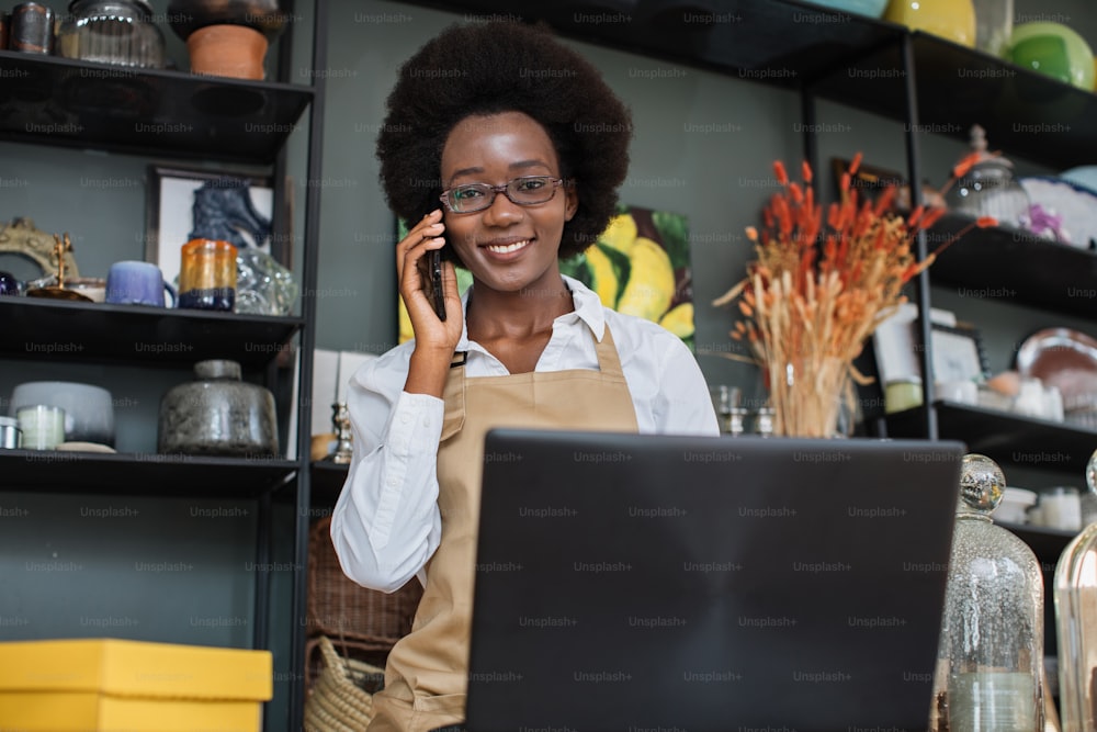 Close up of pretty African saleswoman talking on mobile and typing on laptop at decor shop. Various beautiful decor lying on shelves. Modern gadgets for work.