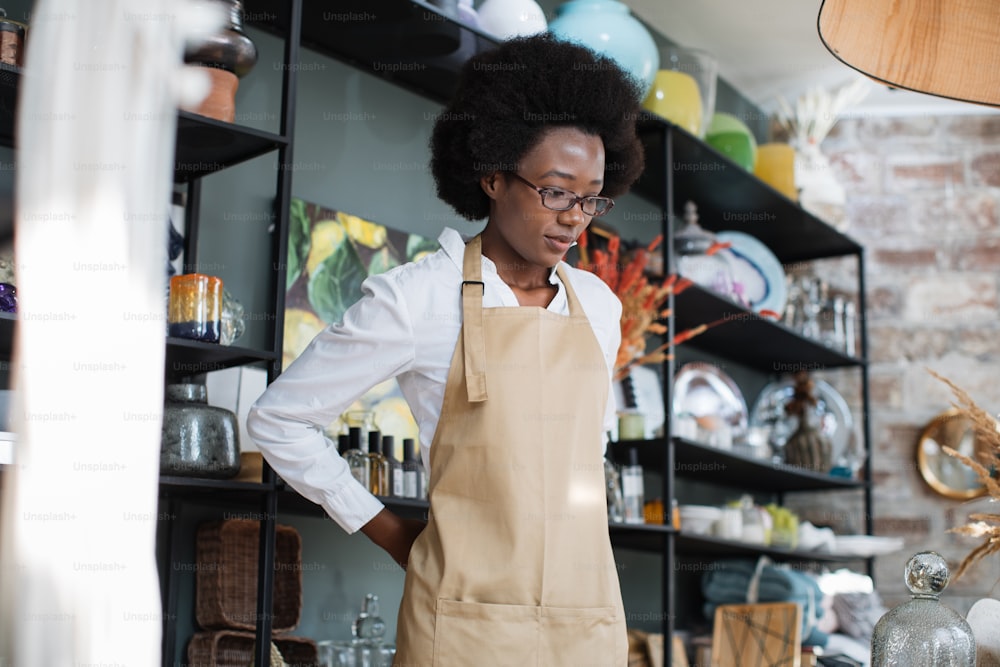 Attraente donna afroamericana che indossa un grembiule beige durante il processo di lavoro nel negozio di decorazioni. Varietà di cose esclusive per la decorazione.