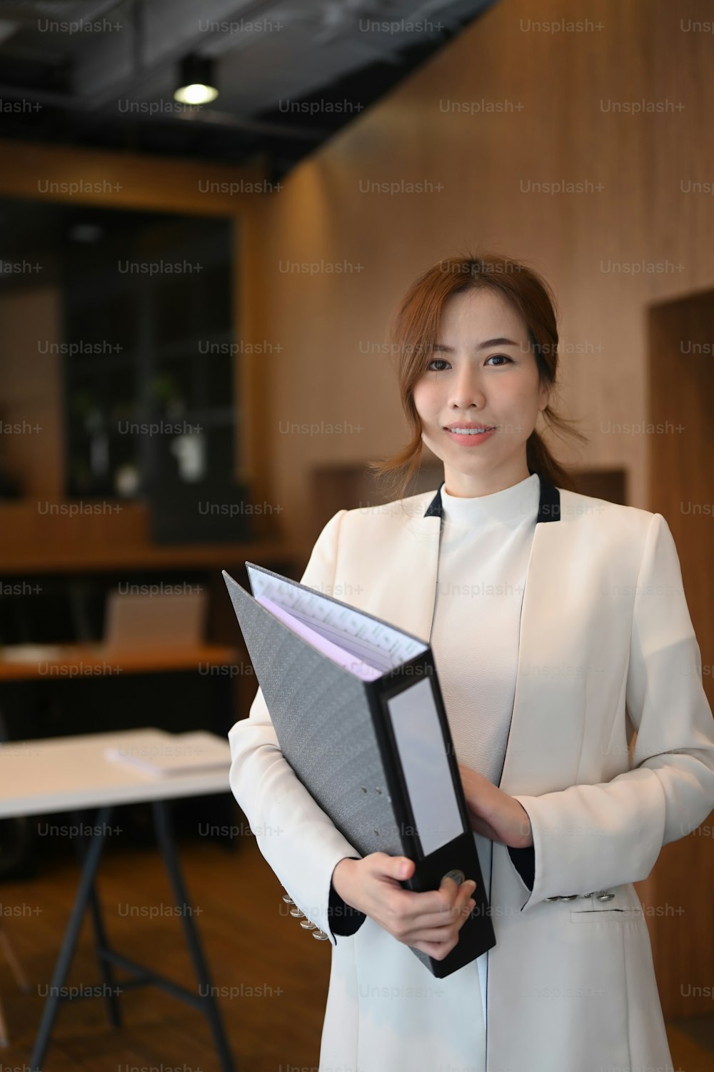Porträt einer weiblichen Führungskraft, die eine Aktenmappe hält und in die Kamera lächelt, während sie an einem modernen Arbeitsplatz steht.
