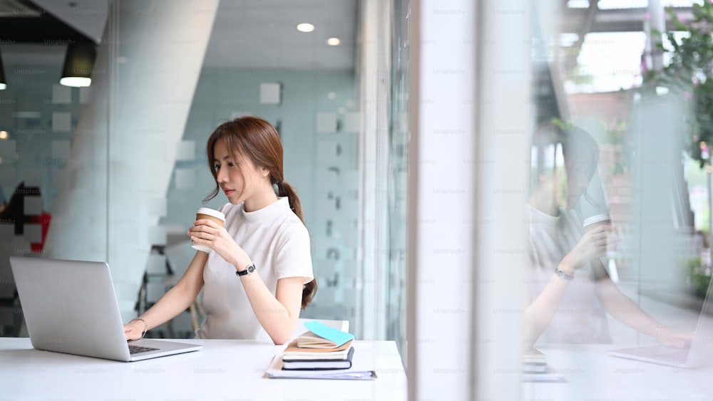 차분한 비즈니스 여성이 창문 근처의 현대적인 직장에 앉아 커피 컵을 들고 노트북 컴퓨터로 작업하고 있다.