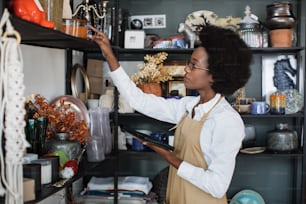 Jolie vendeuse en chemise blanche et tablier beige à carreaux avec tablette moderne au magasin. Femme africaine racontant tous les biens au travail.