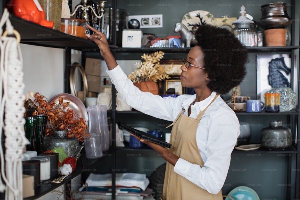 Atractiva vendedora con camisa blanca y delantal beige que comprueba la decoración con una tableta moderna en la tienda. Mujer africana contando todos los bienes en el trabajo.