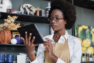 안경과 베이지색 앞치마를 입은 매력적인 아프리카 여성이 장식 상점에서 디지털 태블릿으로 작업하고 있다. 직장에서 재고를 위해 현대 가제트를 사용하는 예쁜 판매원.