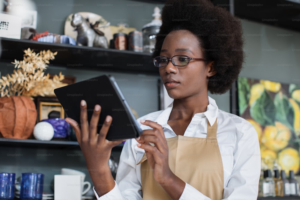 Affascinante donna africana in occhiali da vista e grembiule beige che lavora su una tavoletta digitale presso un negozio di decorazioni. Bella commessa che utilizza gadget moderni per l'inventario al lavoro.