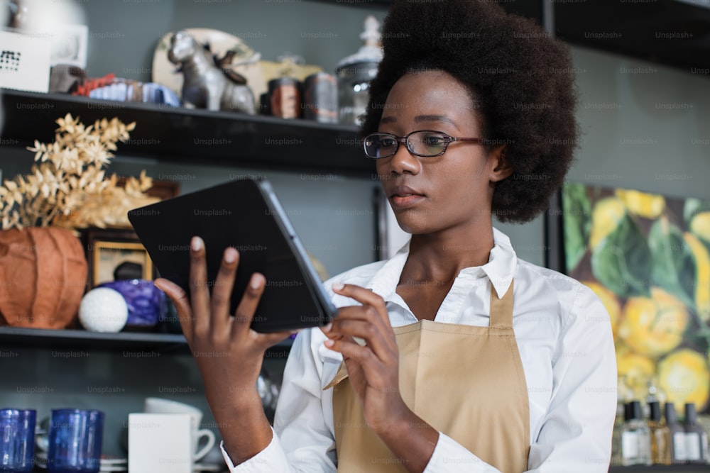 Charming african woman in eyeglasses and beige apron working on digital tablet at decor shop. Pretty saleswoman using modern gadget for inventory at work.