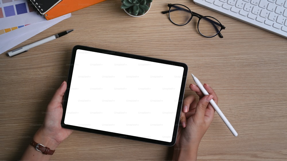 Vue ci-dessus des mains d’une femme tenant une tablette numérique avec écran vide et stylet sur un bureau en bois.