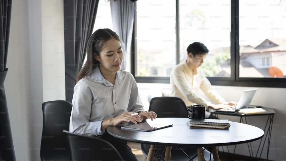 オフィスでデジタルを使用している若いアジアのサラリーマンと彼女の同僚がバックグラウンドで座り込んでいます。