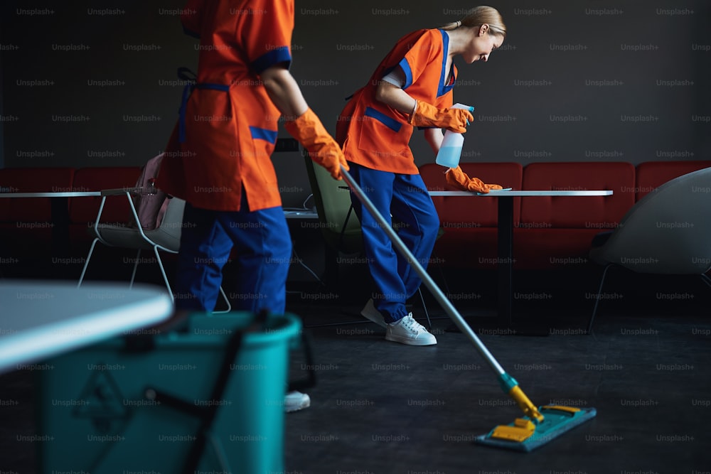 Smiling young blonde Caucasian janitor and her female colleague dressed in uniforms doing the cleaning