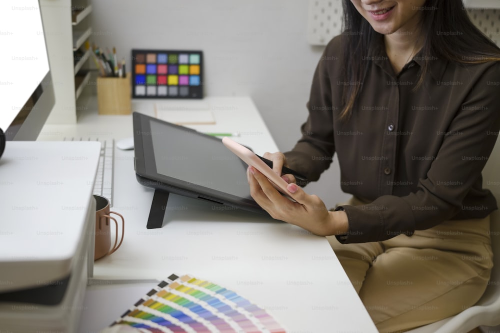 Cropped shot of smiling female graphic designer sitting at her workplace and using mobile phone for texting or massaging with her friends.