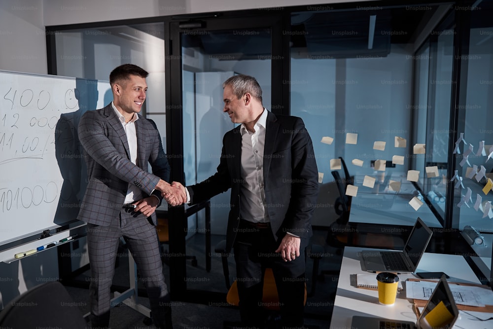 Joyful businessman standing near whiteboard and smiling while making successful deal with business partner