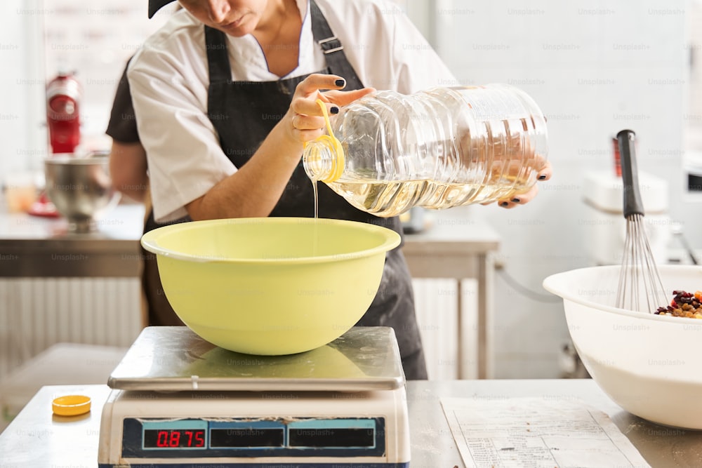 Vista ravvicinata della donna che mette l'olio nel serbatoio e lo pesa mentre prepara l'impasto al tavolo del panificio. Occupazione e concetto alimentare. Immagine