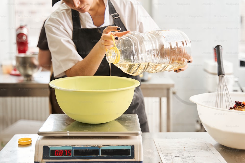 Close up view of the woman putting oil at the tank and weighs it while preparing dough at the table at the bakery. Occupation and food concept. Stock photo