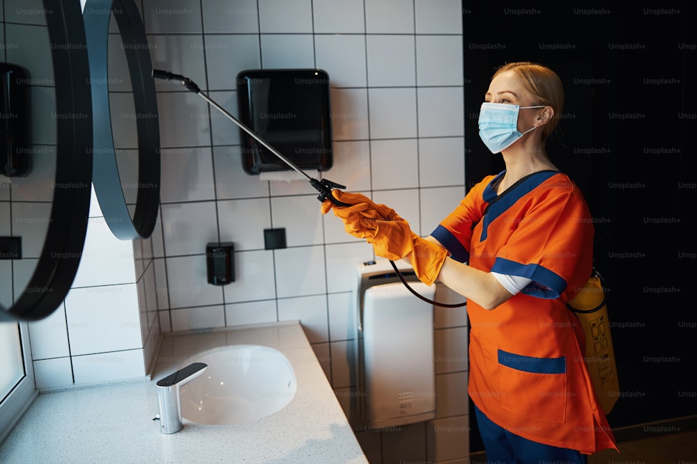 Limpiador calificado con mascarilla desechable y guantes de goma rociando el espejo con un desinfectante