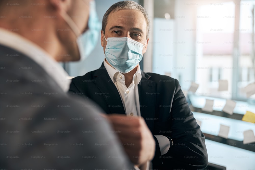 Nahaufnahme eines männlichen Unternehmers, der eine Gesichtsschutzmaske trägt, während er mit einem Geschäftspartner im Büro kommuniziert