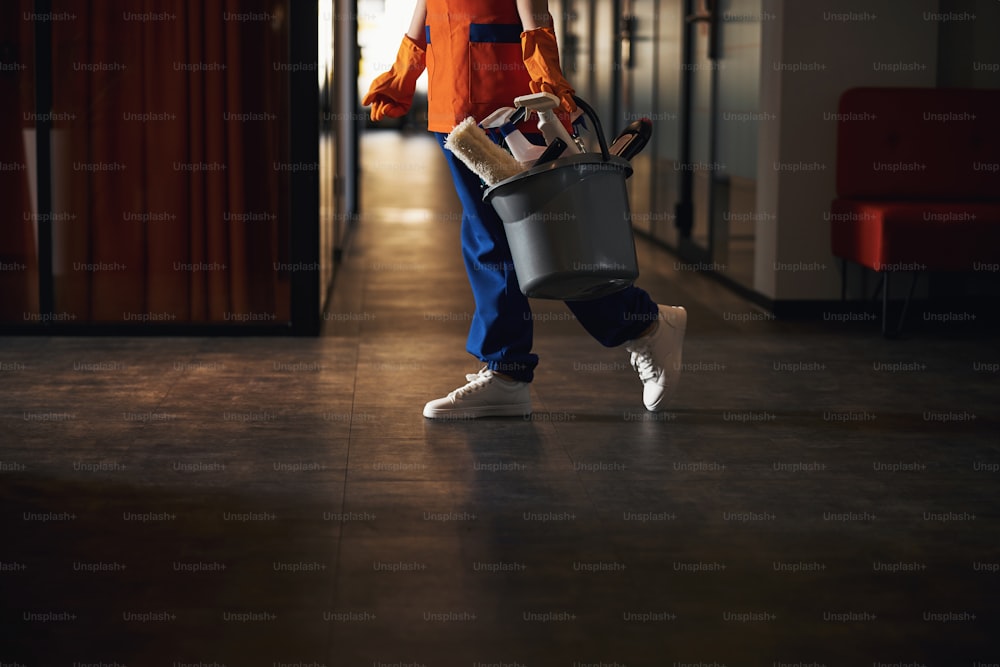 Foto recortada de una mujer con uniforme y guantes de goma que lleva un balde de plástico con suministros de limpieza