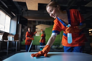 Une femme de ménage pleine d’entrain en gants de caoutchouc et ses collègues en arrière-plan en train de nettoyer le café