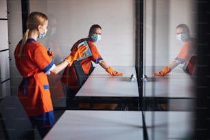 Cleaner in a disposable face mask looking at her blonde female colleague through the glass partition