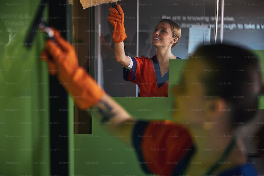 Senhora de limpeza loira sorridente e sua colega lavando as paredes de vidro no escritório