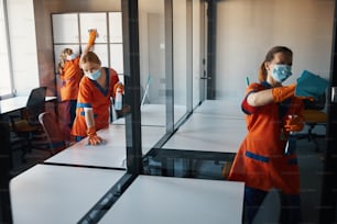 Group of female janitors in rubber gloves and face masks cleaning surfaces in the office