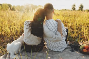 Stilvolles Paar, das sich in sonnigem Licht umarmt und sich mit weißem Hund auf Decke zwischen Gras auf der Sommerwiese entspannt. Sommerurlaub und Picknick. Junge Familie genießt den Sonnenuntergang mit Schweizer Schäferhund Welpe