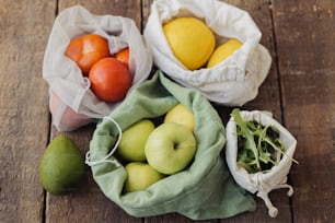 ゼロウェイストショッピング。素朴な木製のテーブルの上に、新鮮なリンゴ、トマト、レモン、アボカド、ルッコラがエココットンの袋に入れられています。オーガニックの果物や野菜を再利用可能な袋に入れたもの。プラスチックフリーの環境に優しい食料品