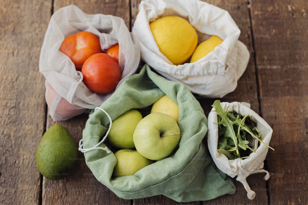 제로 웨이스트 쇼핑. 신선한 사과, 토마토, 레몬, 아보카도, 루꼴라가 소박한 나무 테이블에 담긴 에코 코튼 백에 담겨 있습니다. 재사용 가능한 가방에 담긴 유기농 과일과 채소. 플라스틱 무료 친환경 식료품