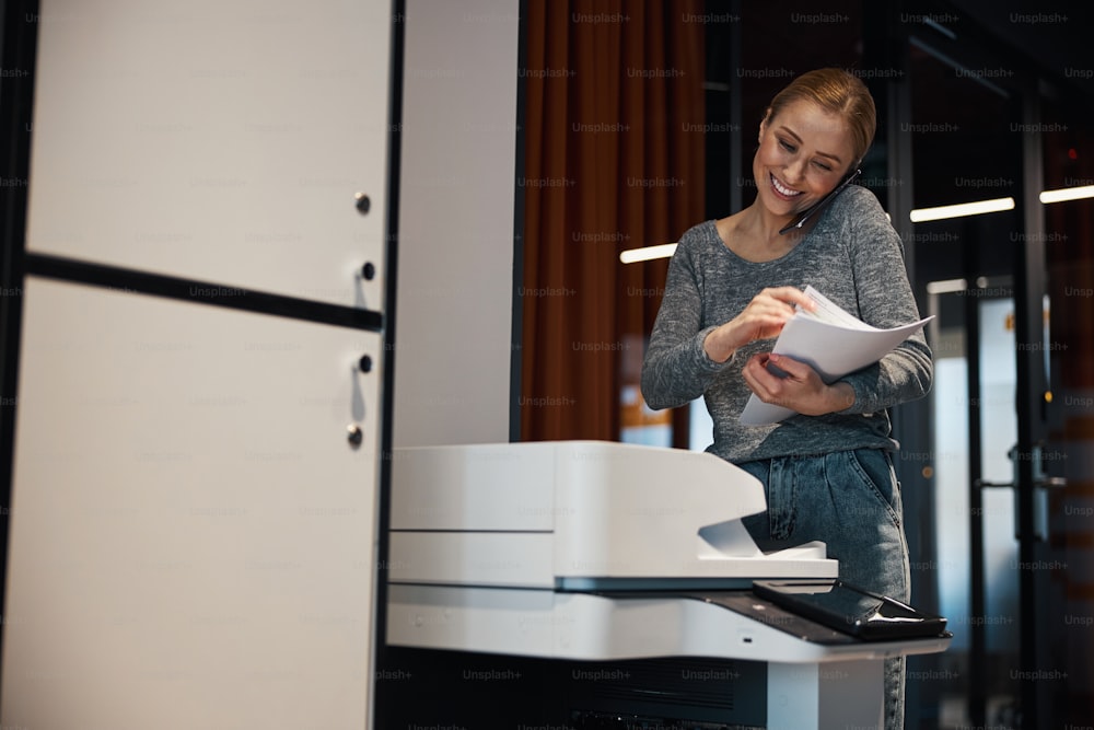 Smiling young blonde woman with a smartphone pressed to the ear standing by the printer