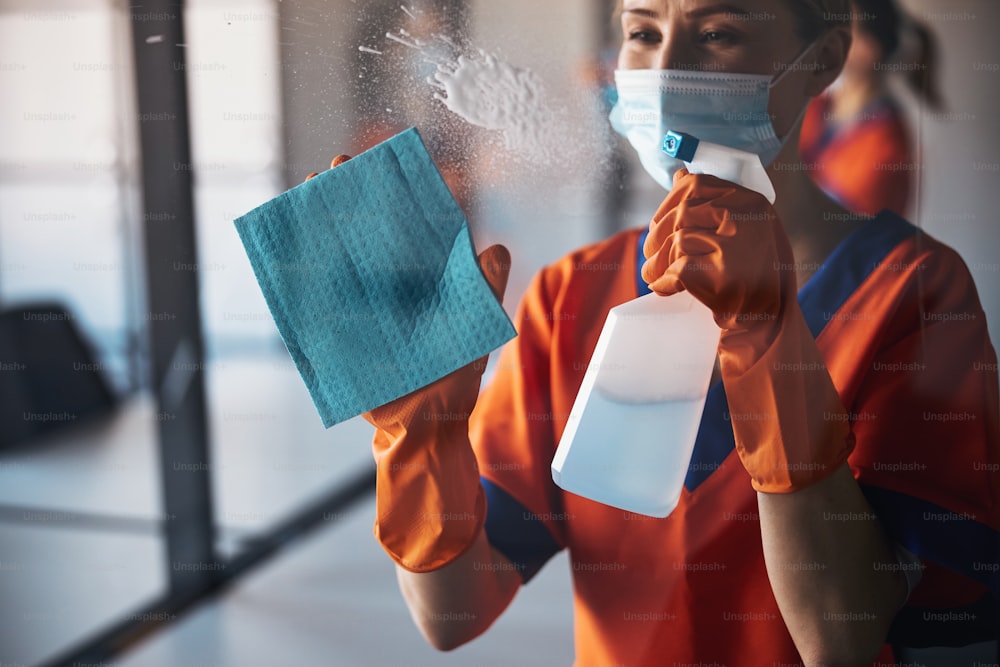 Limpiador profesional con guantes de goma que espolvorea un detergente de una botella de plástico sobre la superficie de vidrio