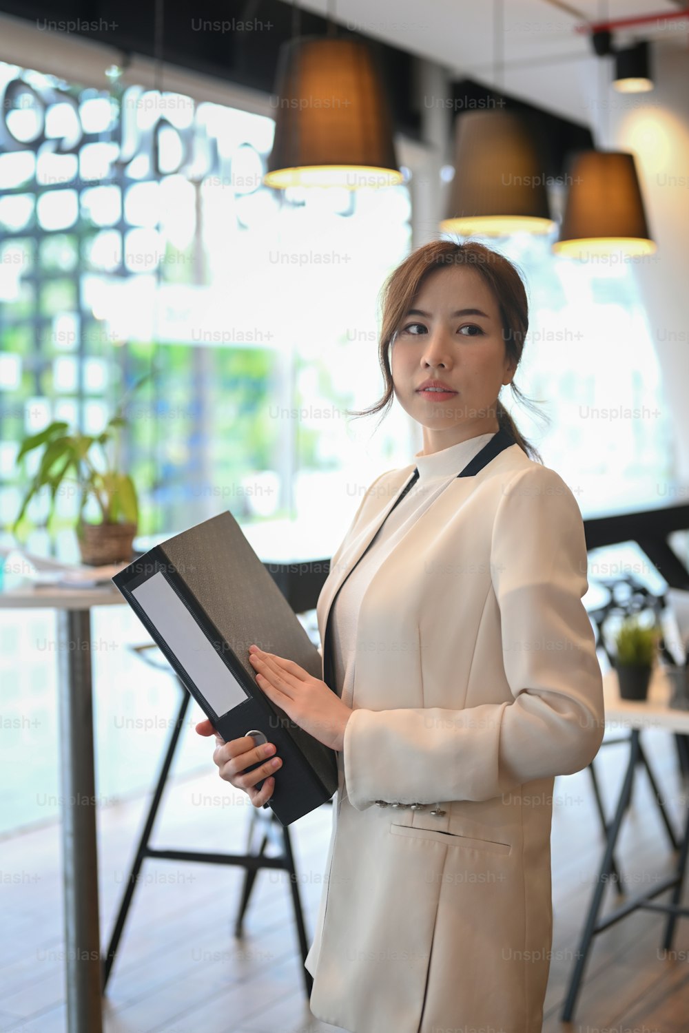 Portrait of confident woman entrepreneur holding file folder and looking away while standing in modern workplace.