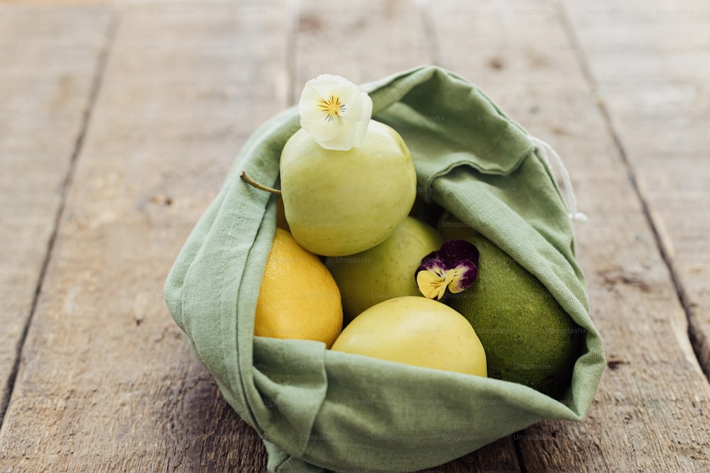 Fresh apples, avocado, lemons in eco cotton bag with flower on rustic wooden table. Zero waste shopping concept. Organic fruits and vegetables in green reusable bag. Eco friendly plastic free