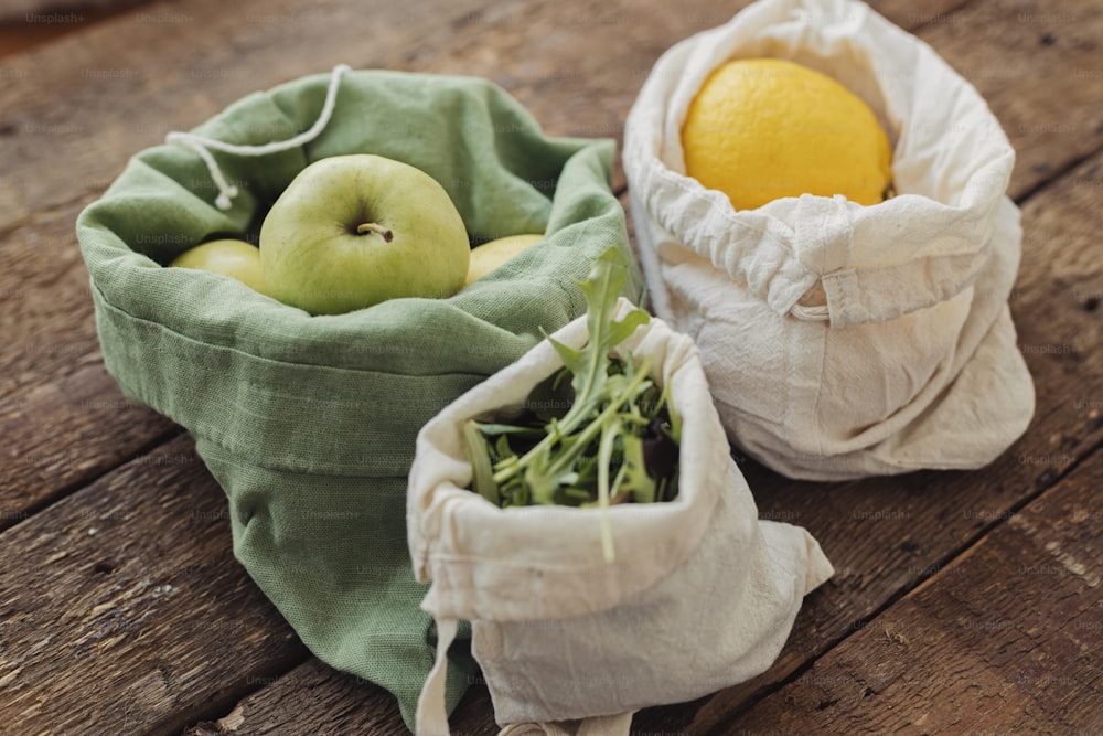 Compras sin residuos. Manzanas frescas, limones y rúcula en bolsas de algodón ecológico sobre mesa rústica de madera. Frutas y verduras ecológicas en bolsas reutilizables. Estilo de vida sostenible. Comestibles sin plástico