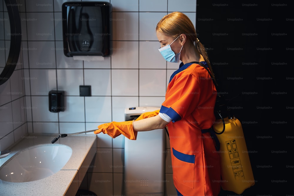 Vista lateral de una limpiadora profesional con mascarilla y guantes de goma desinfectando el baño